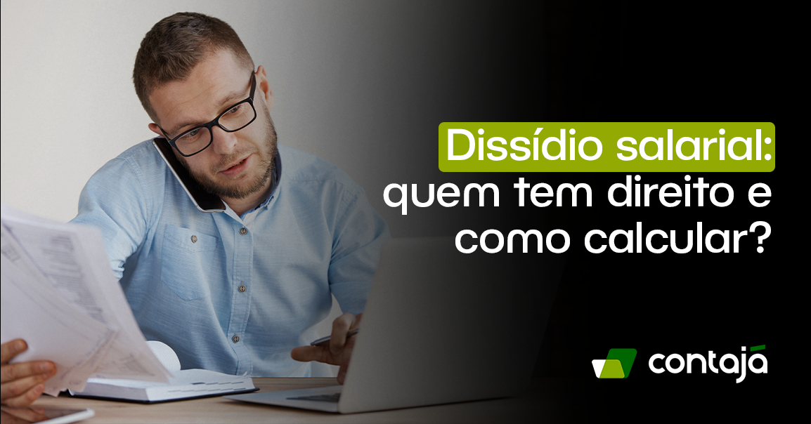 Homem branco com cabelo curto preto, está vestindo uma camisa manga longa azul claro. Ele usa óculos e na imagem está com um celular perto de uma de suas orelhas. Ele está falando no telefone, em uma de suas mãos ele está segurando algumas folhas de papel e com a outra mão ele está digitando no teclado do notebook branco que está na sua frente em uma mesa de escritório. No canto direito da imagem, está o título do artigo: "Dissídio salarial: quem tem direito e como calcular?"