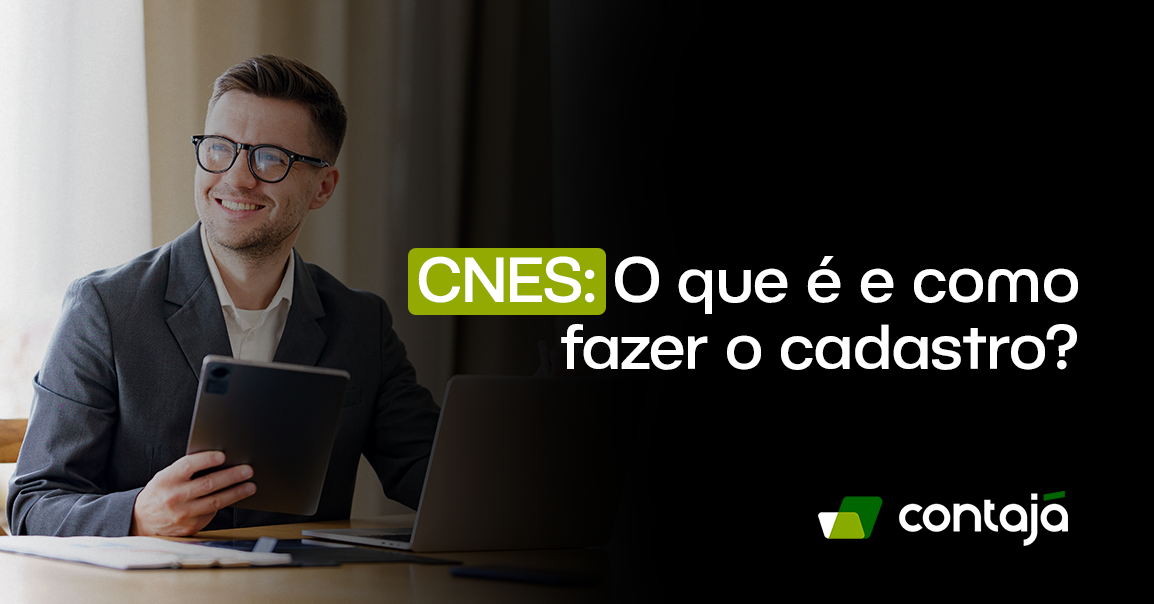Homem branco, vestindo um blazer cinza e uma camisa social branca, ele tem o cabelo preto e está usando óculos de grau. Ele está sorrindo e está olhando para o lado e com uma das mãos ele segura um tablet. Ele está sentado de frente para uma mesa de escritório de madeira. Sobre a mesa tem um notebook cinza. No canto da imagem, está o título do artigo: "CNES: O que é e como fazer o cadastro?"