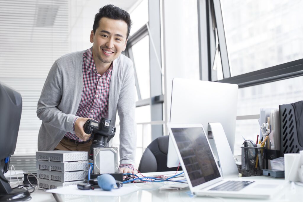 imagem de pessoa sorrindo com câmera na mão e notebook na mesa