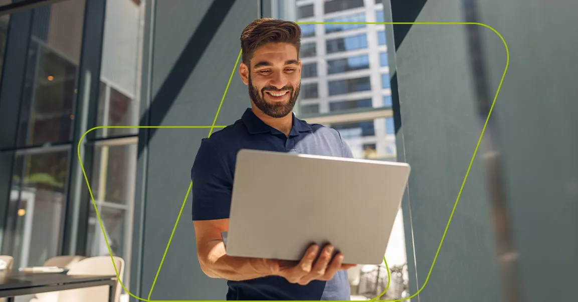 Homem sorrindo com o notebook apoiado no braço, entre prédios envidraçados