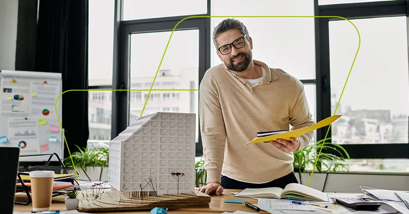 Engenheiro sorrindo na mesa de trabalho, com uma maquete de edifício ao lado e uma pasta de projetos na mão