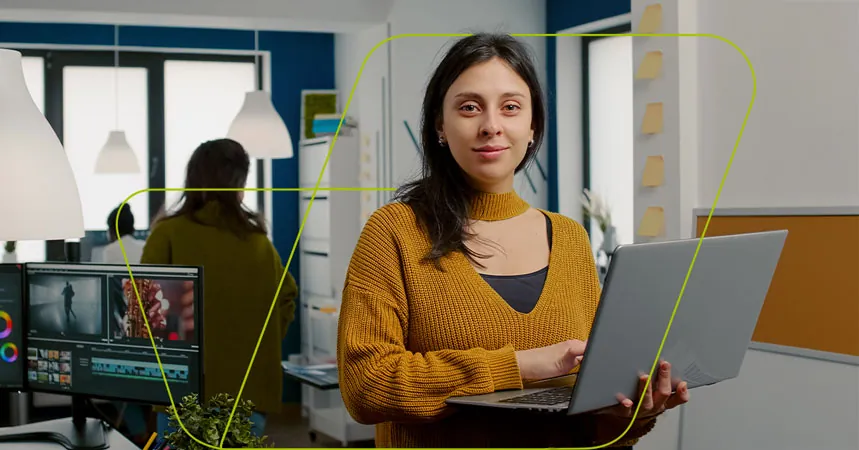 Profissional de marketing em pé numa agência, com o notebook aberto na mão e a estação de edição de vídeo ao fundo