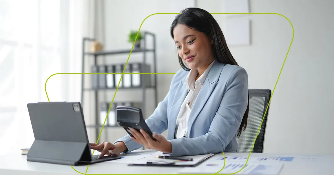 Mulher de blazer claro na mesa de trabalho, conferindo a calculadora com o tablet aberto