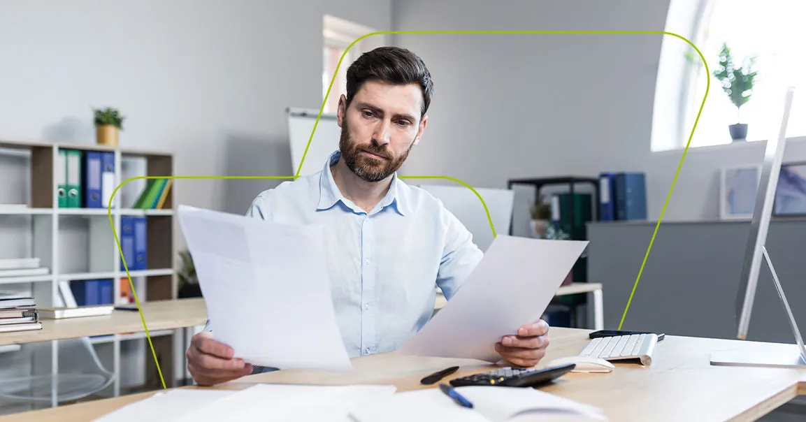 Homem sentado à mesa do escritório comparando dois documentos, com uma calculadora ao lado
