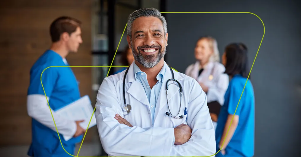 Médico de jaleco branco e estetoscópio sorrindo, com a equipe do hospital ao fundo