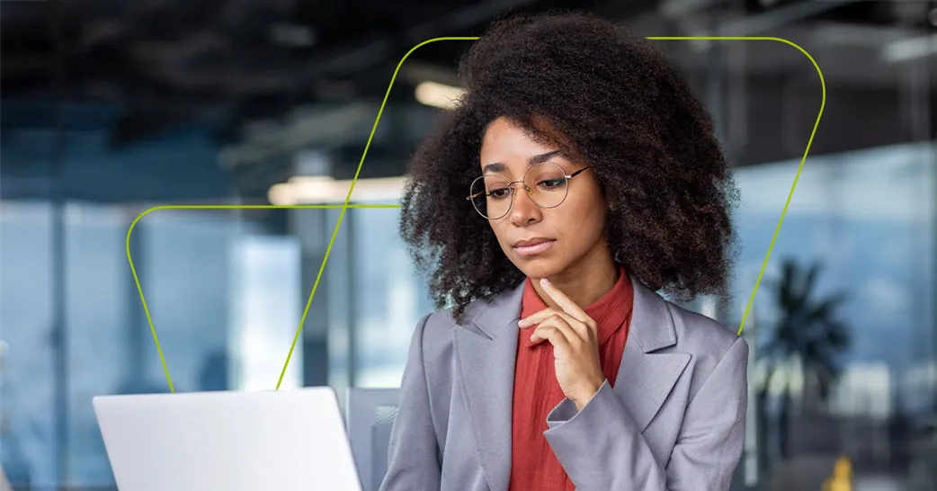 Mulher de óculos e blazer diante do notebook, com a mão no queixo, num escritório envidraçado