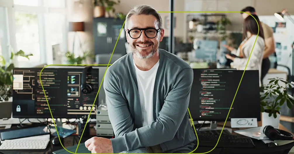 Desenvolvedor de óculos sorrindo entre dois monitores com código aberto, no escritório